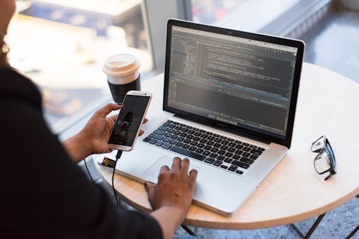 Developer working remotely, coding on a laptop with phone in hand, showcasing modern work culture.